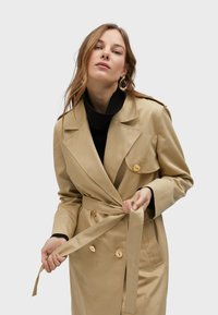 Beige trench coat with a double-breasted design, belted waist, and subtle patterned texture. Features large buttons and a collar.