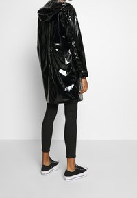 Schwarzer, glänzender Regenmantel mit Kapuze, mittellangem Schnitt und Kordelzug in der Taille, kombiniert mit schwarzen Leggings und lässigen schwarzen Sneakern.