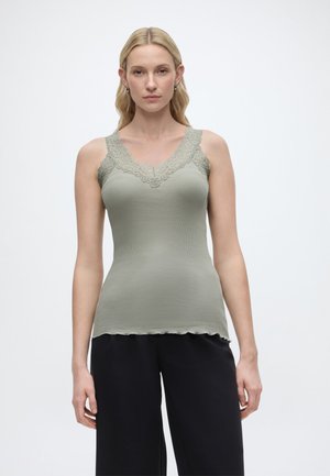 Kvinde med langt blondt hår iført en tætsiddende salviegrøn top med blonder og løse sorte bukser, stående mod en ensfarvet baggrund.