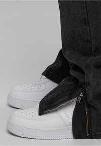 White sneakers with perforated design, worn with dark gray jeans featuring a zippered side opening. Close-up view of footwear and pant detail.