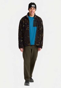 Fleecejacke in Dunkelbraun mit gelben und grünen Mustern, mit Reißverschlusstaschen, kombiniert mit einem blauen T-Shirt und olivgrünen Cargohosen. Schwarze Mütze auf dem Kopf.