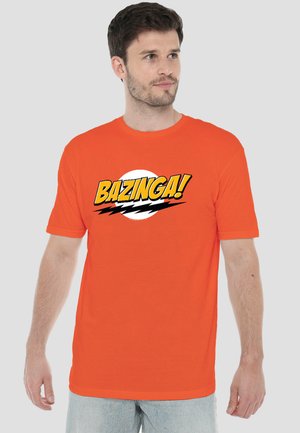 Man wearing orange T-shirt with "BAZINGA!" text and black lightning bolt graphic, looking to his left.