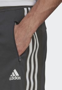 Pantalones cortos atléticos grises con un bolsillo con cremallera, que presentan tres franjas verticales blancas a lo largo del costado y un logotipo bordado en blanco.