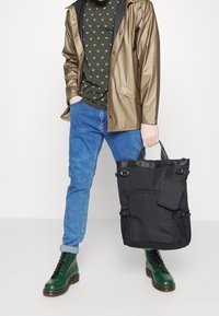 Svart tygkasse med justerbara remmar, texturerat tyg, sidofickor och en liten fastsatt väska. Modellen bär en brun jacka och gröna stövlar.