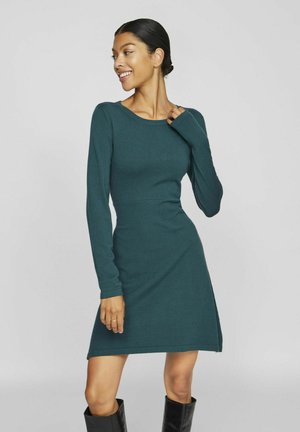 Robe ajustée à manches longues en tissu tricoté teal foncé. Dotée d'un col rond et d'une fente sur le côté. Portée avec des bottes noires jusqu'aux genoux.
