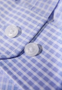 Błękitno-biała kraciasta tkanina z dwoma białymi guzikami o teksturowanym wzorze, przedstawiająca zbliżenie kołnierzyka i listwy koszuli.