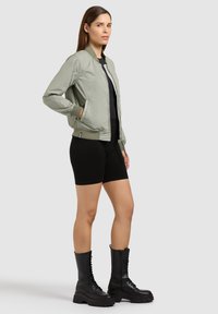 Femme debout, les mains dans les poches, portant une veste bomber vert clair, un short noir et des bottes de combat noires sur un fond uni.