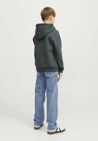 Felpa verde con cappuccio e finitura testurizzata, abbinata a jeans azzurri chiari. La persona indossa sneakers bianche con dettagli neri.