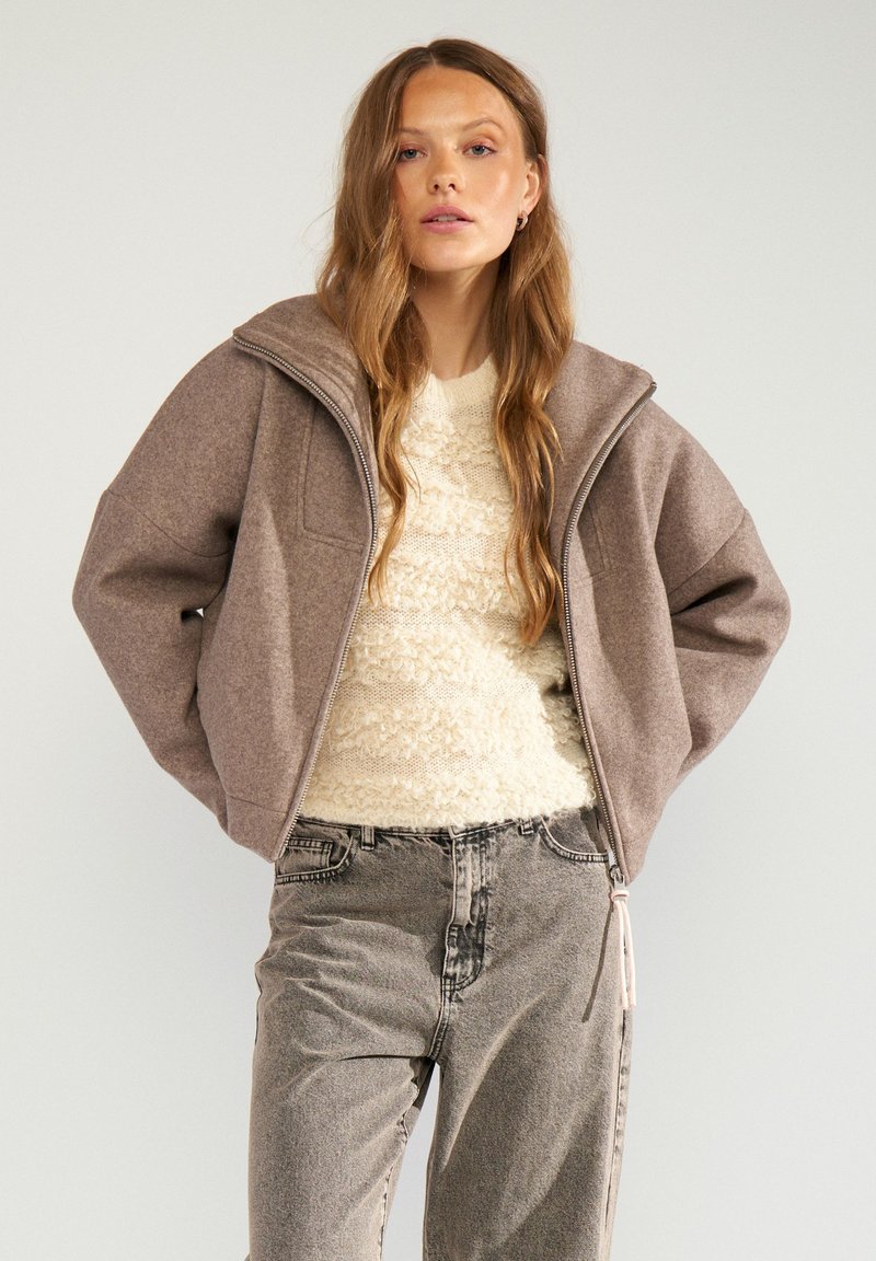 Femme aux longs cheveux portant une veste marron zippée, un pull crème texturé et un jean gris délavé, debout devant un fond uni.