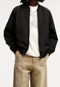 Svart bomberjacka med dragkedja och ribbade ärmslut, lagd över en vit t-shirt med blå tryck, tillsammans med beige vida byxor.