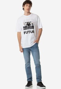 Camiseta blanca de algodón con un estampado gráfico que presenta la palabra "FUTUR" y una imagen en blanco y negro. Llevada con jeans de mezclilla azul claro y zapatillas negras.