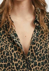Žena v hnědočerné košili s leopardím potiskem na knoflíky, rozepnut�é v horní části, která odhaluje dekolt a část hrudníku.