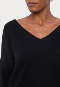 Woman wearing a black V-neck sweater, showing neckline and collarbones, with a silver earring visible on one ear.