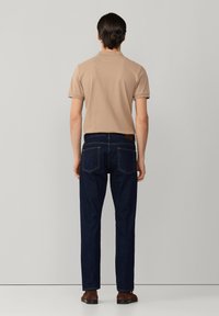 Muž v béžovej polokošeli s krátkym rukávom a tmavomodrých džínsach stojaci čelom k svetlosivej stene, viditeľné hnedé topánky.