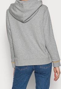 Grijze hoodie van zacht materiaal, met een capuchon, ribgebreide manchetten en een gouden accent aan de mouwzoom. Draagt met blauwe jeans.