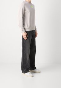 Lichtgrijze trui met geribbelde manchetten en tailleband, met een klein groen logo. Gecombineerd met donkergrijze wijde jeans en witte sneakers.