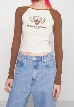 Persoon met felrood haar, gekleed in een crèmekleurige en bruine cropped top met lange mouwen en het logo "Après Club", en een lichtblauwe high-waisted spijkerbroek.