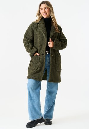 Olive green, fluffy faux fur coat with large front pockets, button closure, and a relaxed fit. Worn over a black top and blue jeans.
