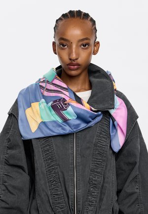 La sciarpa di seta presenta un design astratto colorato con forme geometriche in blu, rosa e giallo, drappeggiata attorno al collo di una modella sopra un giacca grigia.