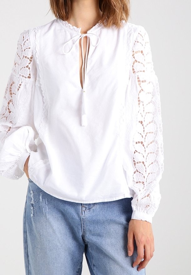 Blouse blanche avec détails en dentelle sur les manches longues, coupe ample et encolure en trou de serrure avec un lien. Portée avec un jean bleu clair déchiré.