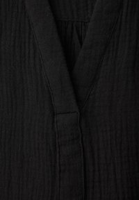 Lähikuva mustasta teksturoidusta kankaasta, jossa näkyy V-pääntie, taitettu kaulus ja ommellut saumedetaljit.