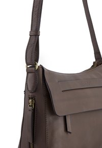 Brązowa skórzana torebka o wyraźnej formie, z klapką jako zapięciem, mosiężnymi akcentami i teksturowanym wykończeniem. Widoczna kieszeń boczna na zamek.