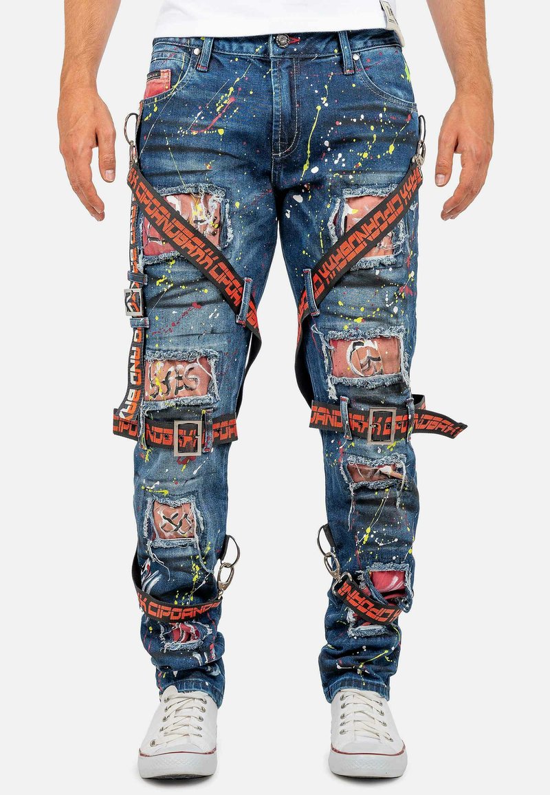 Jean en denim bleu avec un design usé, des éclaboussures colorées, de multiples poches à patch et de larges sangles avec des accents de texte rouge et noir.