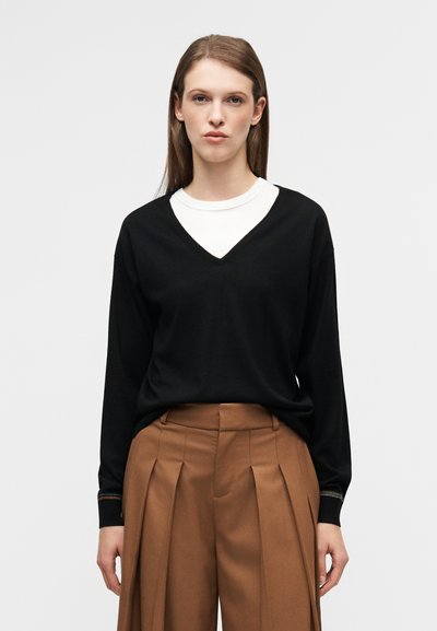 Μαύρο πουλόβερ με λαιμόκοψη σε σχήμα V και μακριά μανίκια, φορεμένο πάνω από λευκό μπλουζάκι με στρογγυλή λαιμόκοψη. Το μοντέλο φοράει καφέ πλισέ παντελόνι και στέκεται μπροστά σε ουδέτερο φόντο.