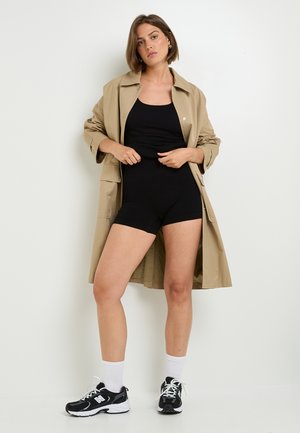 Trench coat beige sopra un bodysuit nero, abbinato a sneakers bianche e nere. La modella è in piedi di fronte a uno sfondo bianco, mostrando i dettagli dell'outfit.