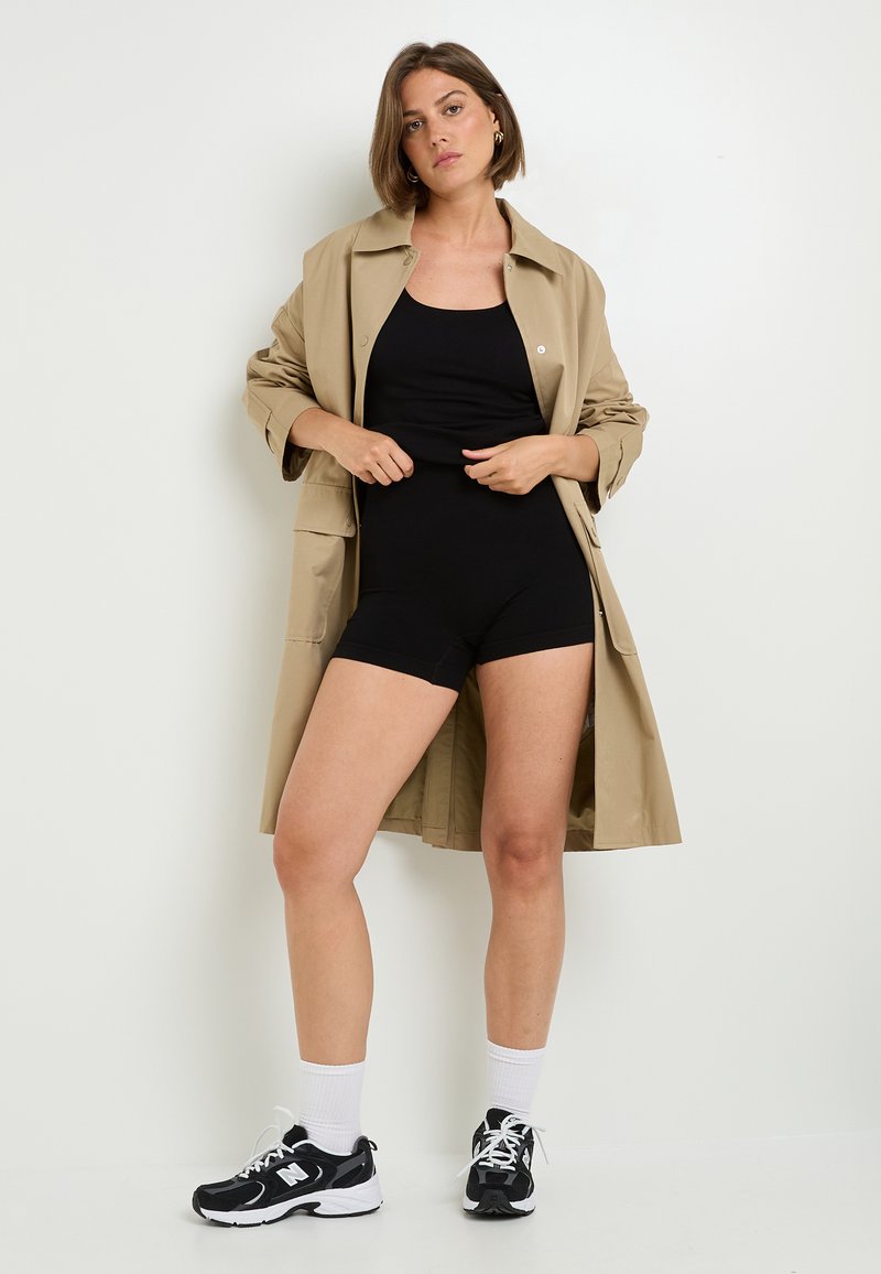 Trench coat beige sopra un bodysuit nero, abbinato a sneakers bianche e nere. La modella è in piedi di fronte a uno sfondo bianco, mostrando i dettagli dell'outfit.