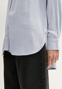 Camicia azzurra chiaro con colletto e orlo curvo, maniche lunghe e tessuto testurizzato. Abbinata a jeans scuri, con un'etichetta visibile di lato.