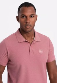 Rosa pikétröja med krage, korta ärmar och en liten broderad logotyp på vänster bröst. Tillverkad av strukturerad tyg.