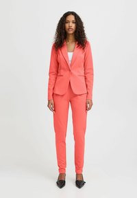 Korallenfarbener Blazer mit Slim Fit, einreihiger Knopfschluss und Reverskragen, kombiniert mit passenden tapered Pants. Strukturierter Stoff, polierte schwarze High Heels.