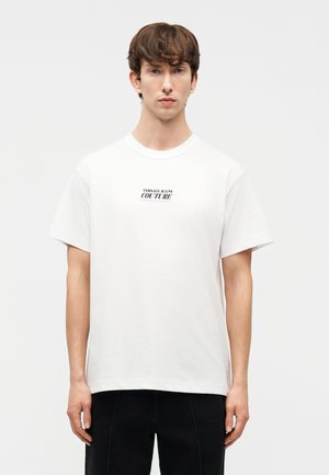 Jonge man met kort bruin haar draagt een eenvoudig wit T-shirt met de opdruk "Versace Jeans Couture" centraal op de borst, gecombineerd met een zwarte broek.