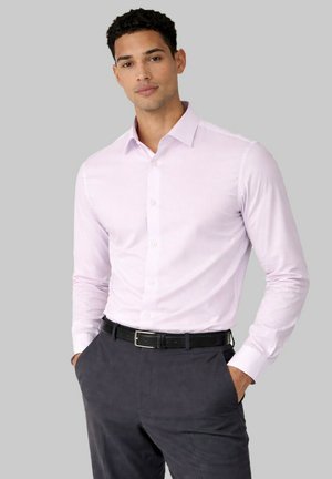 Homme portant une chemise rose clair boutonnée rentrée dans un pantalon gris foncé avec ceinture noire, les mains dans les poches, sur un fond gris uni.