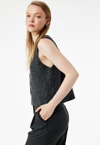 Grå ærmeløs strikvest med ribbet tekstur og åbne sider, båret over mørke bukser. Simpelt design uden synlige lukninger eller mønstre.