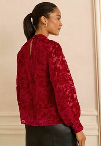 Röd blommig spetsblus med långa puffärmar, detalj med nyckelhål i ryggen och strukturerad tyg. Buren med en svart lädeskjol.