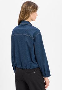 Dark blue, long-sleeve top with a collar, elasticized hem, and visible stitching details. The fabric appears soft with a slight texture.