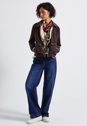 Donna con una giacca marrone scuro, sciarpa a fantasia, jeans blu a gamba larga e sneaker bianche in piedi con le mani in tasca che guarda a destra.