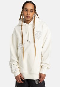 Cream-colored oversized hoodie with a kangaroo pocket, drawstring hood, and graphic designs on both sleeves in gray. Soft texture, relaxed fit.