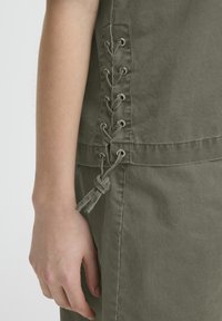 Bras gauche à côté d'un haut en tissu vert olive avec un détail lacé sur le côté noué en un nœud près de la hanche.