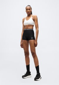 Γυμνασμένη γυναίκα σε όρθια στάση, φοράει λευκό αθλητικό σουτιέν, μαύρα συμπιεστικά σορτς Under Armour, μαύρες κάλτσες και μαύρα αθλητικά παπούτσια.