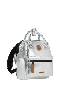 Silver backpack with a glossy finish, dual handles, adjustable black strap, front zip pocket, and a tan logo patch. Compact and structured design.