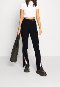 Zwarte, rekbare broek met zijsplitten, gecombineerd met een witte cropped top met een halve rits. Model houdt een grijze tas vast en draagt dikke schoenen.
