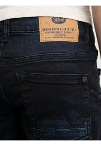 Vaqueros de mezclilla azul oscuro con un parche de cuero que muestra el logo de la marca, costuras visibles y un bolsillo trasero con detalles decorativos en la costura.