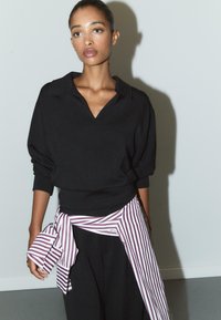 Sweatshirt nero oversize con scollo a V e spalle scese, abbinato a una giacca a strisce bianca e bordeaux legata sopra pantaloni neri.
