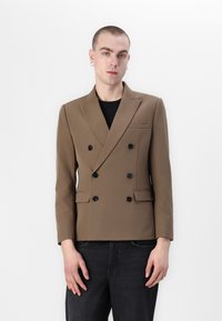 Blazer marrón de doble botonadura hecho de tela texturizada, con botones negros, solapas con muescas y un bolsillo en el pecho. Llevado con jeans oscuros.