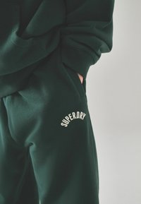 Ciemnozielony sweter i spodnie dresowe z miękkiego materiału, z białym haftowanym logo "SUPERDRY" na spodniach. Luźny fason z kieszeniami.