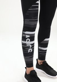 Mustat leggingsit abstrakteilla harmailla ja valkoisilla raidoilla, joissa on suuri valkoinen logo. Valmistettu venyvästä materiaalista, suunniteltu aktiiviseen käyttöön.