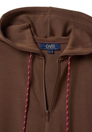 Sudadera con capucha marrón con cremallera, cordones rojos y blancos, y una etiqueta azul en el interior del cuello que dice "OVS" y muestra la información de talla.